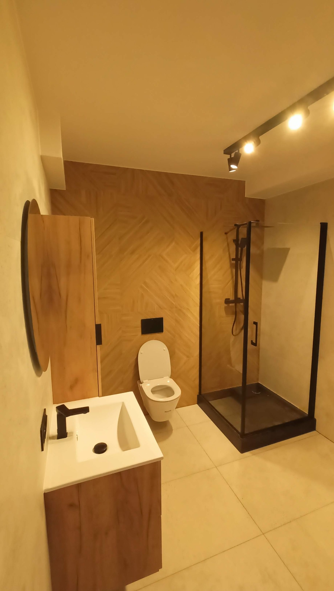 Nowoczesna łazienka z kabiną prysznicową typu walk-in z czarnymi ramami, umywalką z szafką w odcieniach drewna i toaletą. Ściana wykończona panelami imitującymi drewno.