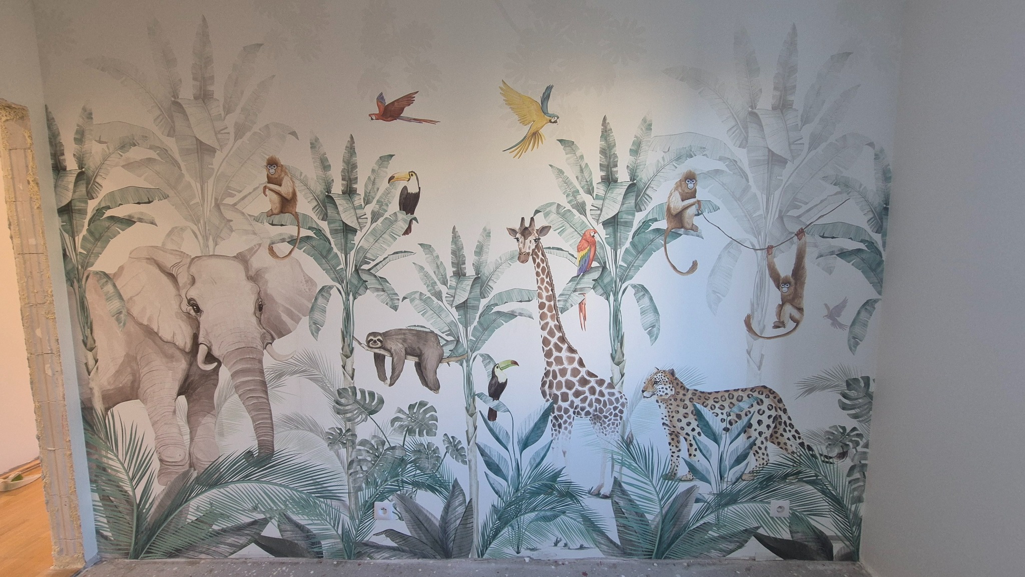 Ściana z fototapetą w motywy dżungli: słoń, żyrafa, małpy, leniwiec i leopard wśród tropikalnych roślin. Widoczne gniazdka elektryczne i fragment surowej ściany.