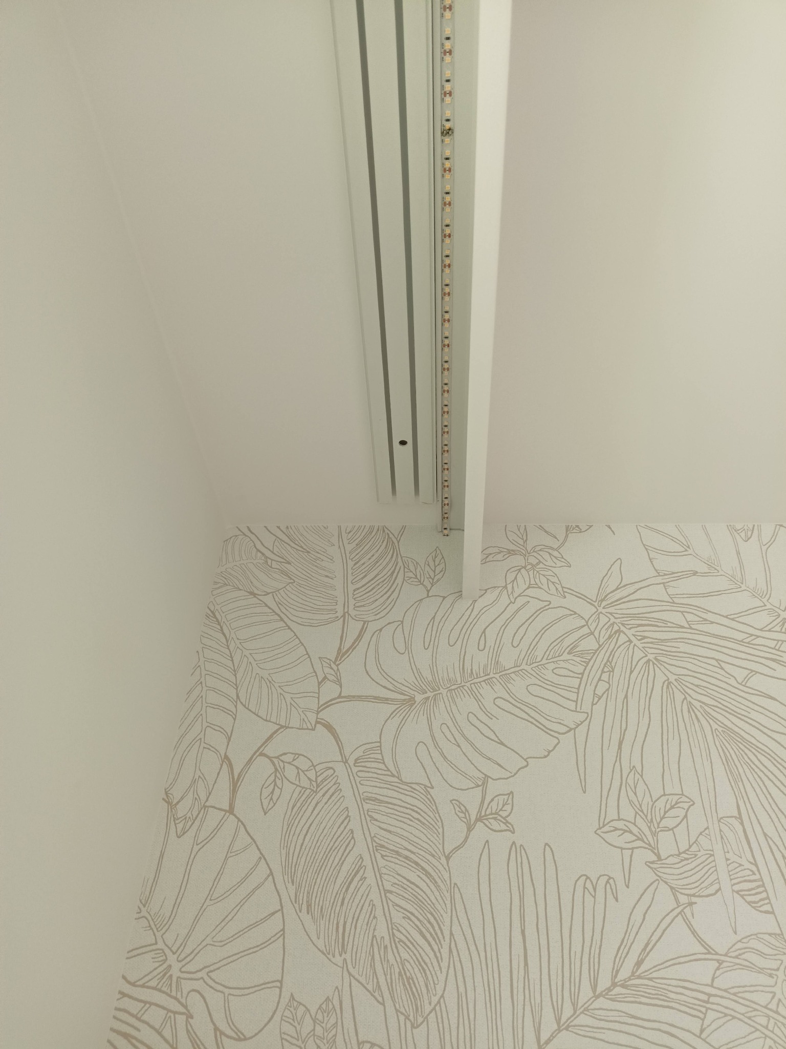 Biała listwa sufitowa z oświetleniem LED, zamontowana przy ścianie z tapetą w motywy roślinne. Ujęcie z dołu, widoczny fragment sufitu i ściany.