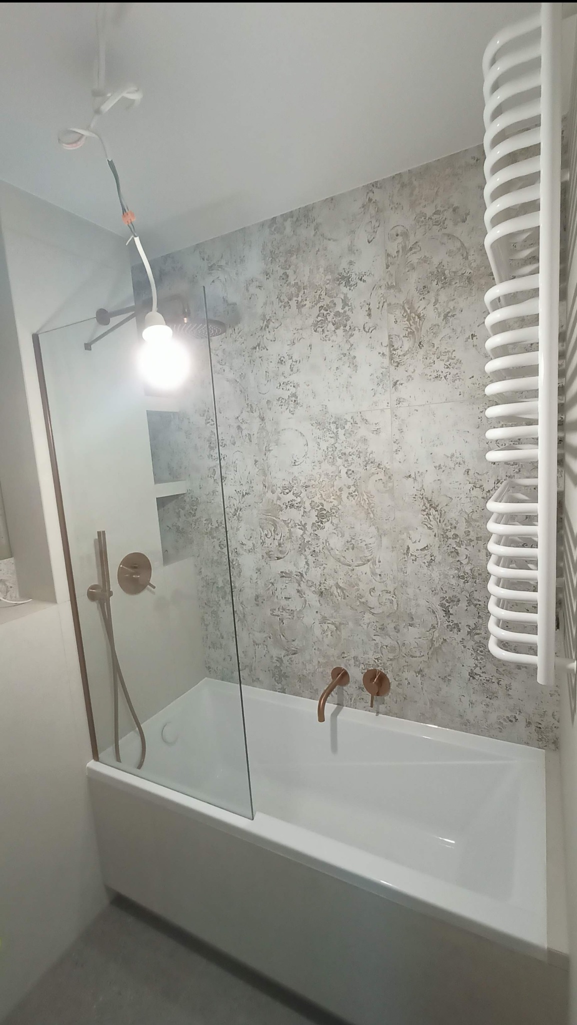 Wykończona łazienka z wanną, szklanym parawanem i grzejnikiem drabinkowym. Na ścianie dekoracyjne płytki w odcieniach szarości. Bateria w kolorze miedzi.