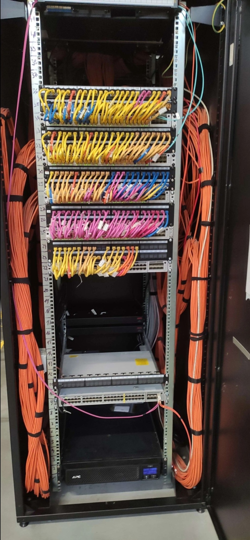 Szafa serwerowa z uporządkowanymi kablami patchcord, oznaczonymi kolorami i numerami, oraz pomarańczowymi kablami światłowodowymi po bokach.