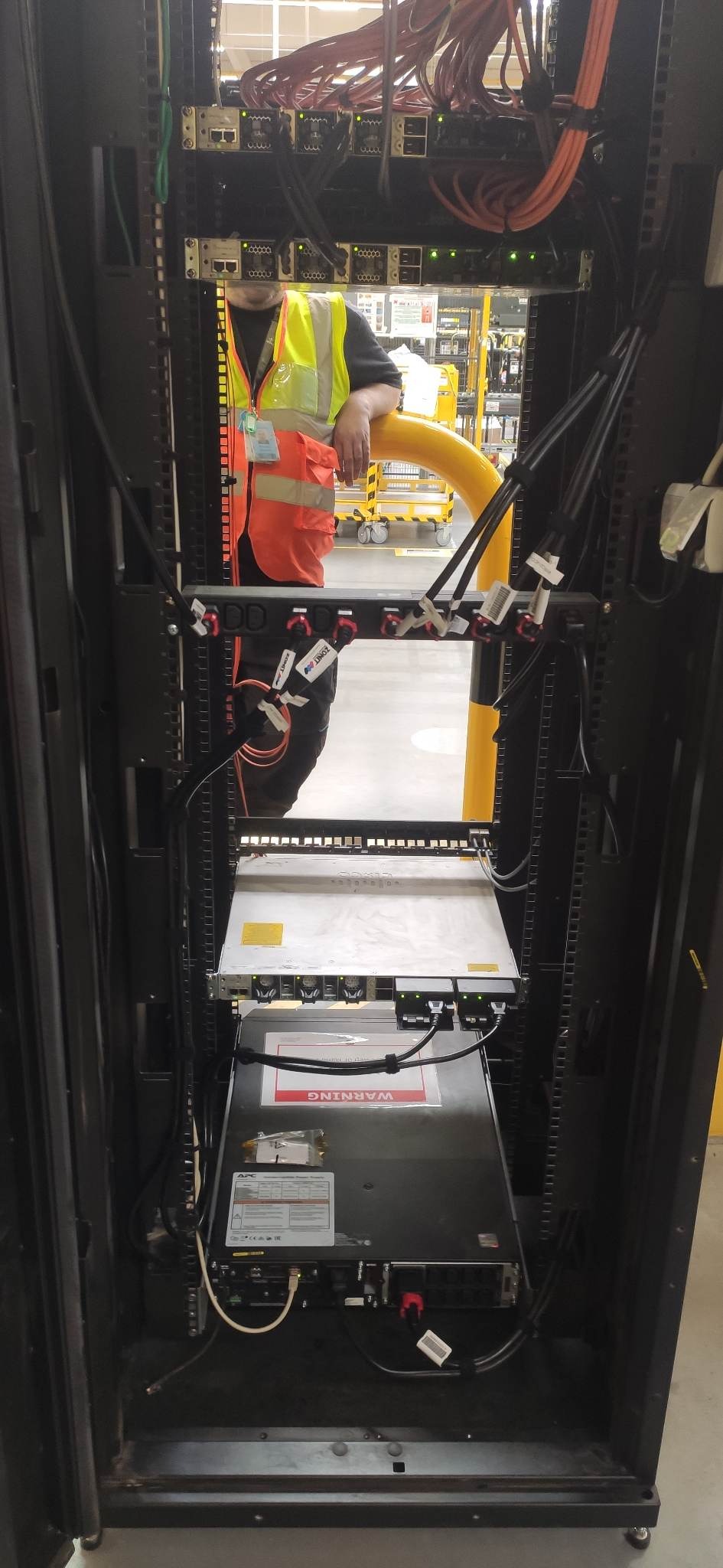 Serwerownia z pracownikiem w kamizelce odblaskowej, widoczne kable, przełączniki i zasilacze. Elementy sieci komputerowej w szafie rack.