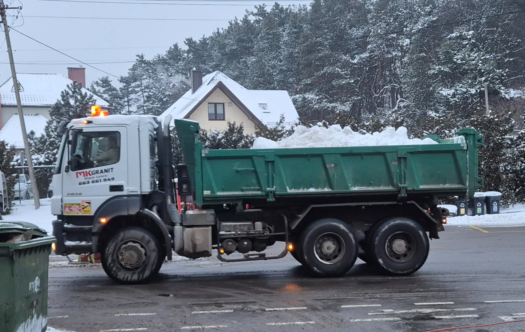 Ciężarówka z zieloną skrzynią wypełnioną śniegiem na tle zimowego krajobrazu z domami i drzewami. Widoczny numer telefonu firmy na kabinie.