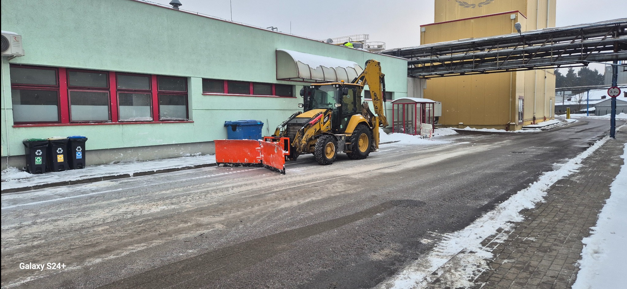 Żółta koparko-ładowarka z pomarańczowym pługiem odśnieżnym na drodze w zimowy dzień, budynek przemysłowy w tle. Ślady opon na jezdni.