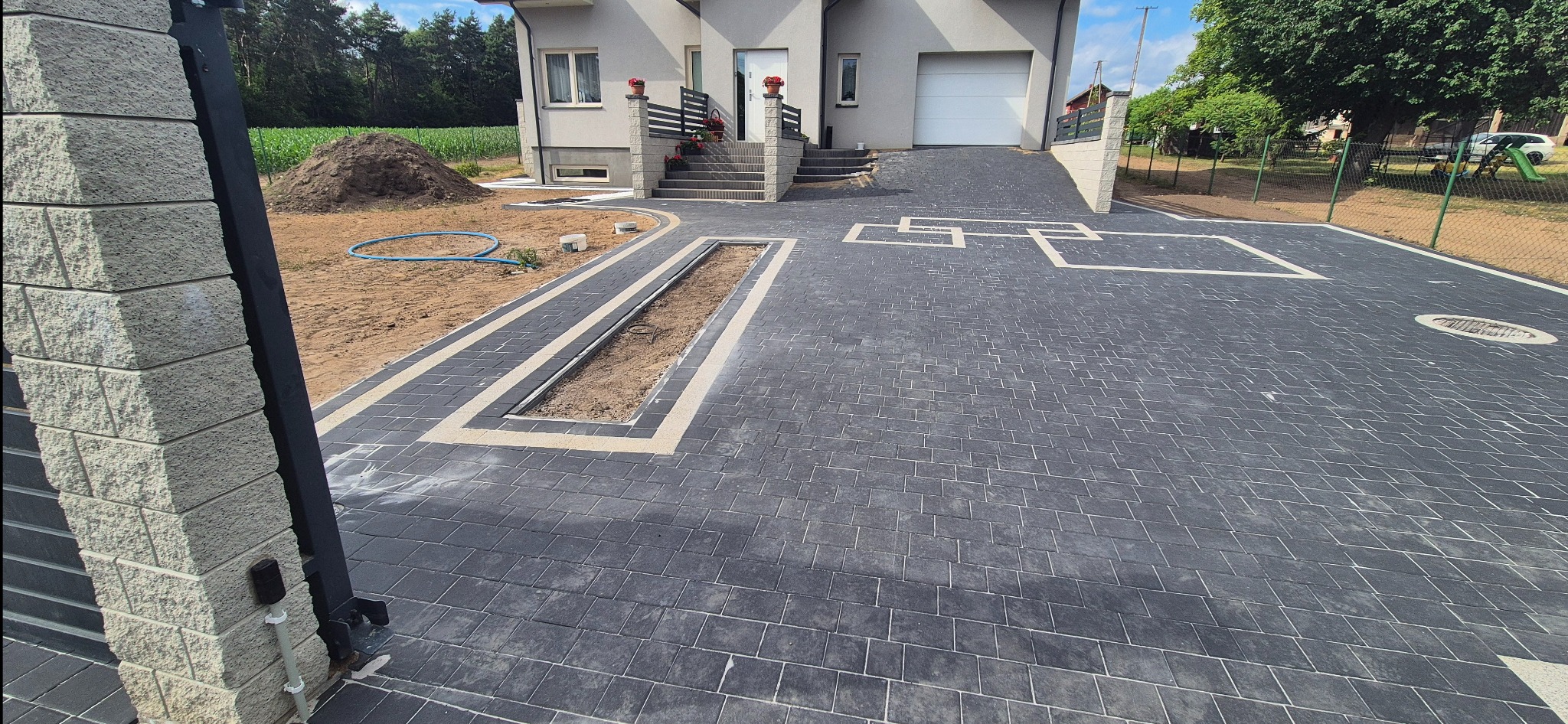 Nowoczesna, szara kostka brukowa z jasnymi obramowaniami na podjeździe przed domem. Widoczny fragment trawnika i brama wjazdowa. Częściowo wykonane odwodnienie liniowe.