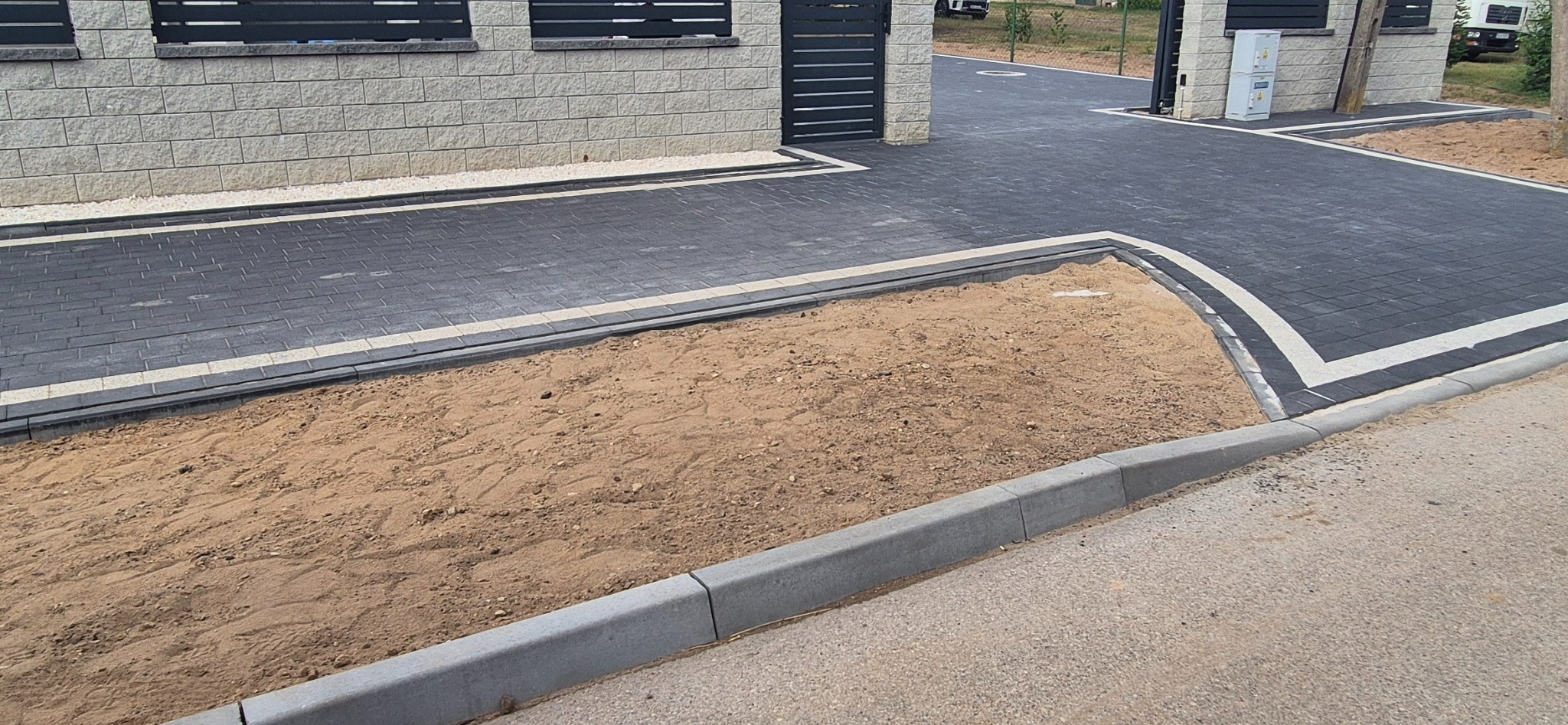 Ułożona, grafitowa kostka brukowa z białymi akcentami, obrzeża i krawężniki, fragment piasku przed posesją. Widok na bramę wjazdową i budynek.