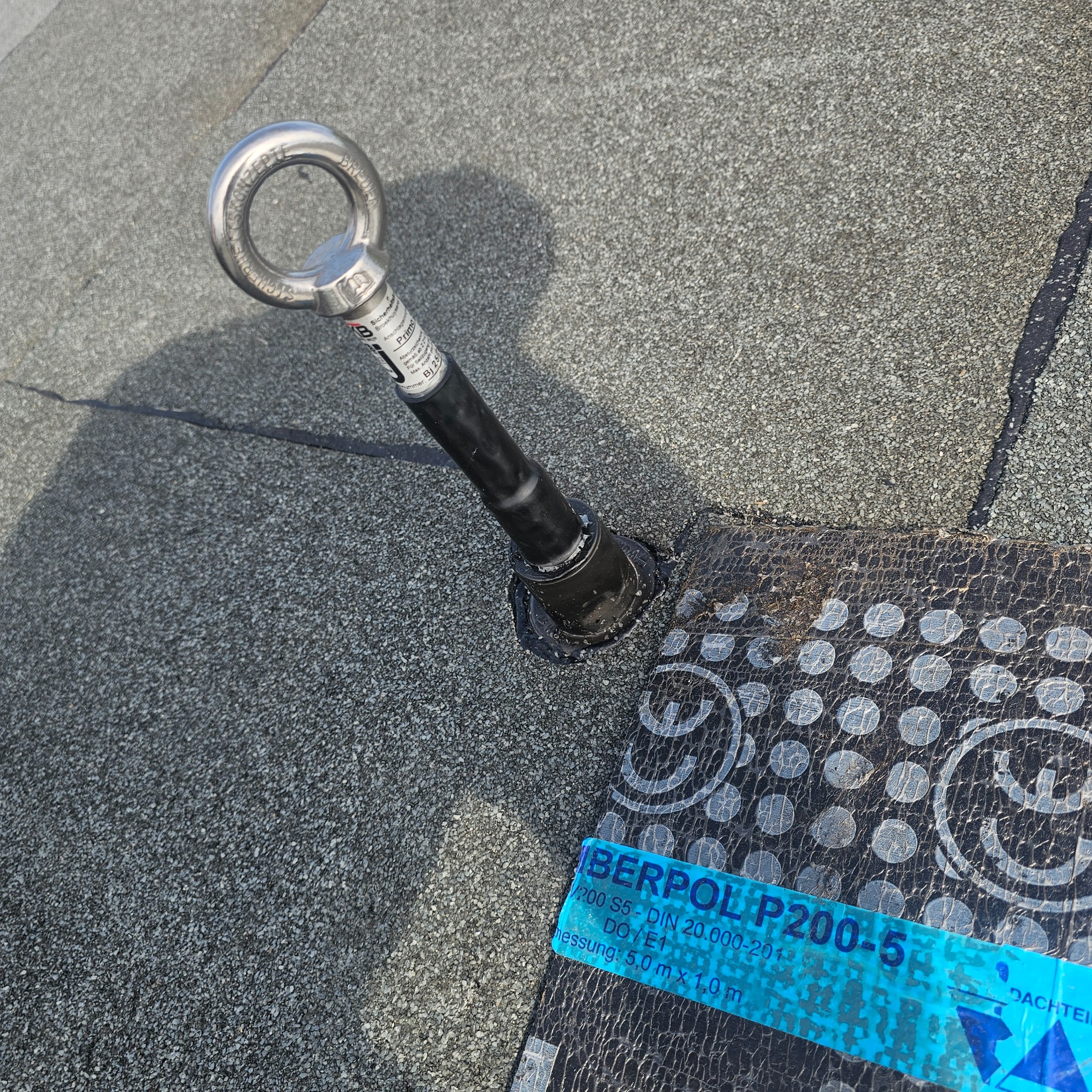 Metalowy uchwyt asekuracyjny zamocowany na papie dachowej z posypką, zabezpieczony czarnym uszczelniaczem. Widoczna etykieta produktu Berpol P200-5.