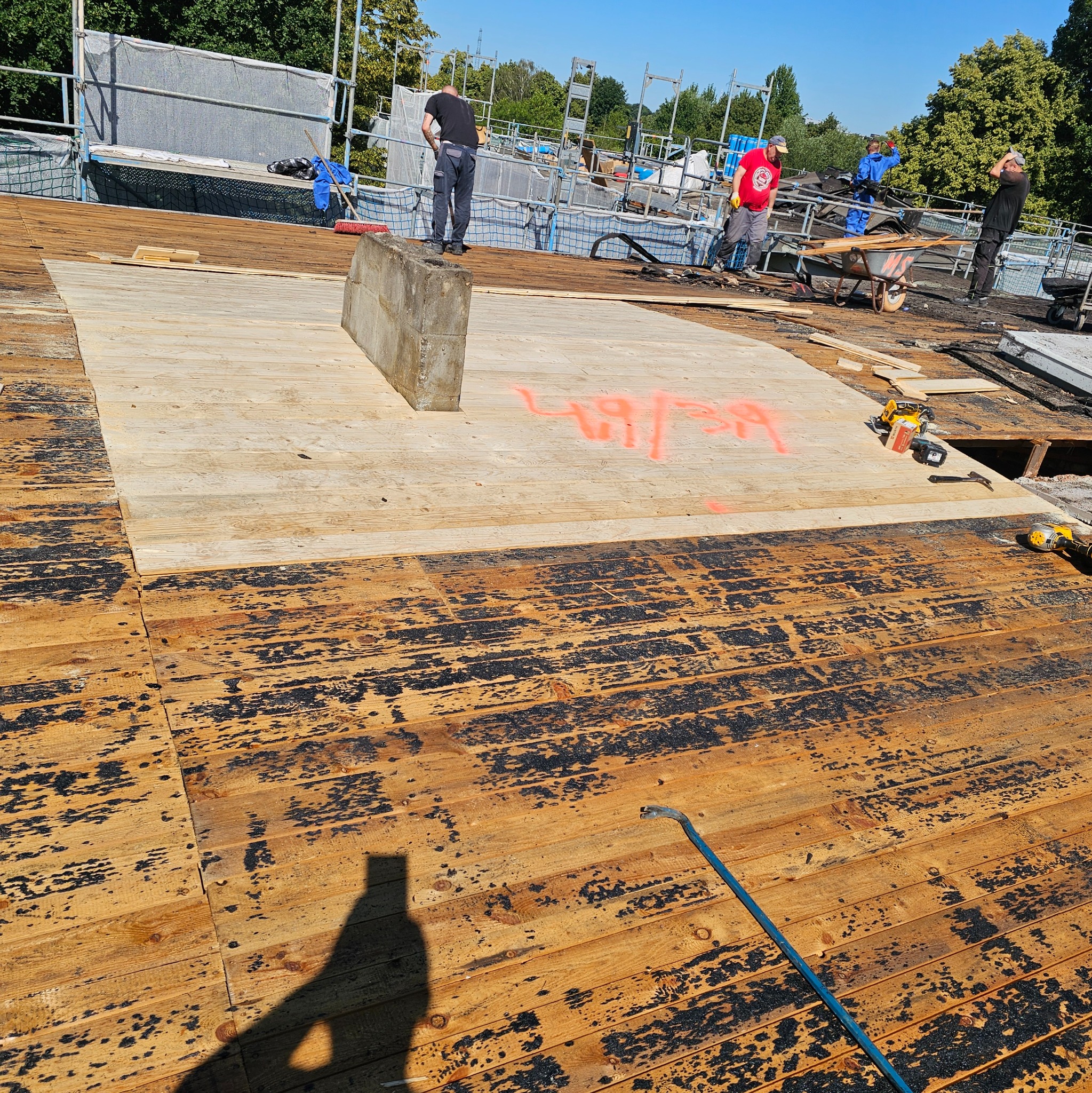 Wymiana fragmentu poszycia dachu z desek. Widoczne narzędzia i ekipa remontowa. Część desek z czarnym nalotem, fragment z nowymi deskami z czerwonymi oznaczeniami.