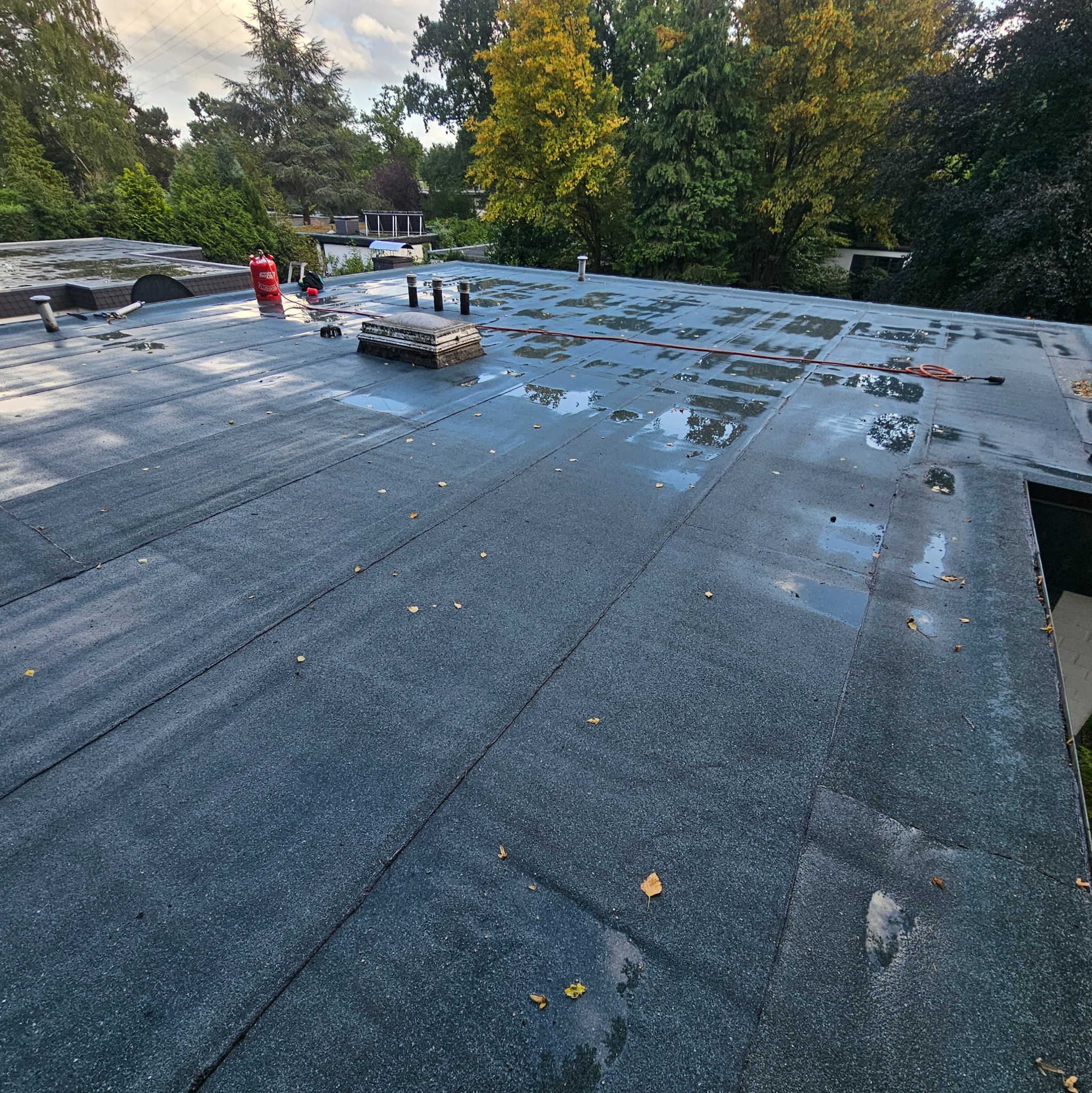 Płaski dach pokryty papą z widocznymi narzędziami dekarskimi, butlą gazową i kałużami po deszczu. W tle drzewa i zabudowania.