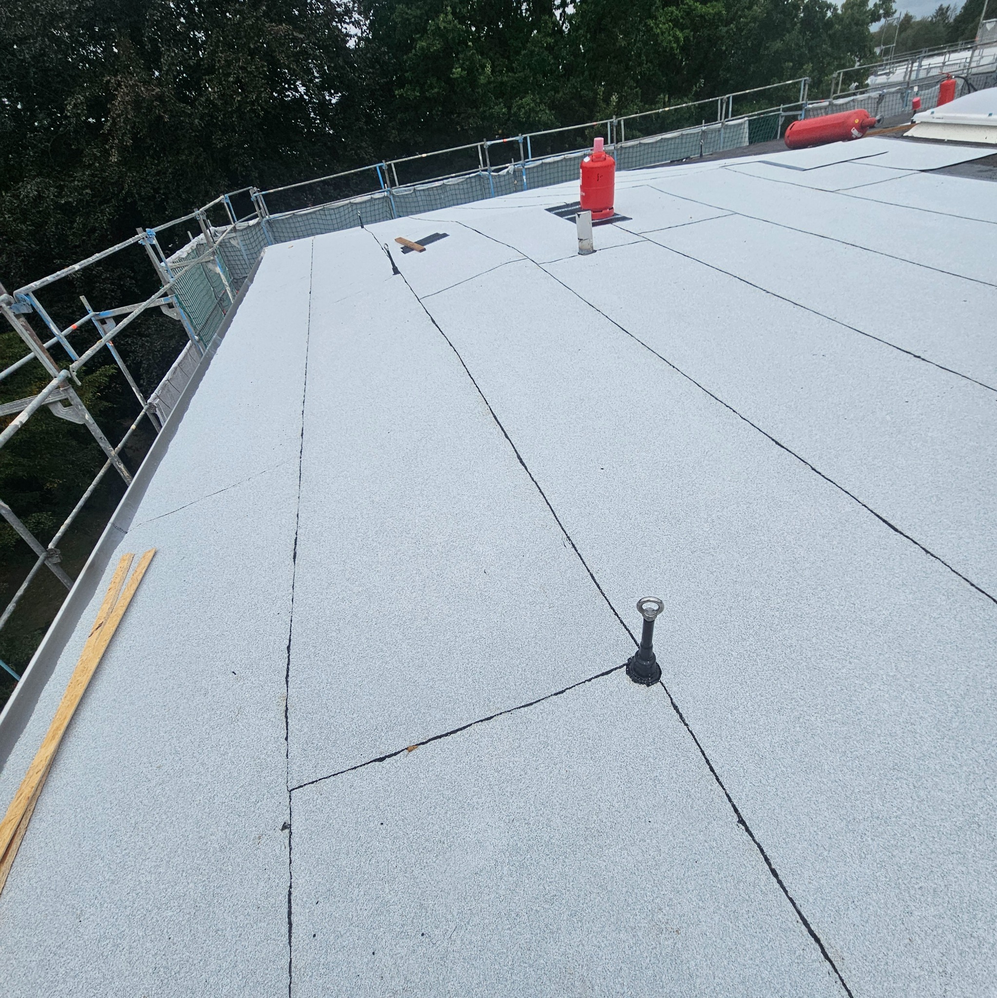 Jasnoszary dach płaski z widocznymi łączeniami papy, zabezpieczeniami i elementami instalacji. Widok z góry, lekki kąt.