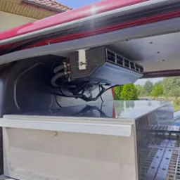 Zamontowany system klimatyzacji w przestrzeni ładunkowej pojazdu, widoczne przewody i kratka wentylacyjna, na tle zabudowy i otoczenia zewnętrznego.