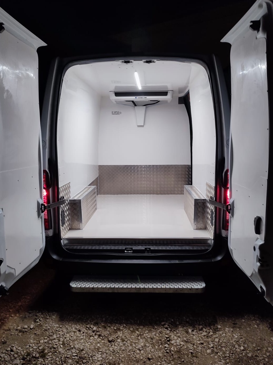 Wnętrze furgonetki chłodni z otwartymi drzwiami, widoczny agregat chłodniczy, aluminiowa podłoga i ściany, oświetlenie wewnętrzne.