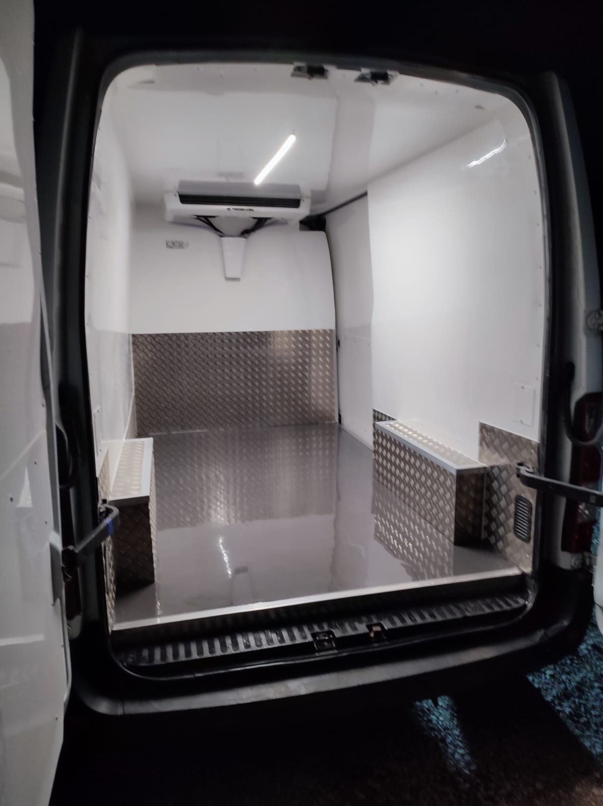 Wnętrze furgonetki chłodniczej z zamontowaną klimatyzacją, widok z tyłu przez otwarte drzwi, na podłodze i ścianach panele z blachy ryflowanej.