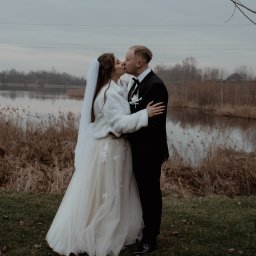 FOCUS4 PRODUCTION Klaudia Karczmarz - Pocałunek pary młodej w plenerze nad jeziorem, panna młoda w białym futerku i sukni z koronkowymi aplikacjami, pan młody w czarnym garniturze.