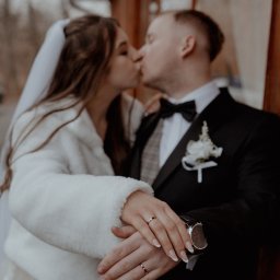 FOCUS4 PRODUCTION Klaudia Karczmarz - Pocałunek nowożeńców ze złotymi obrączkami w Świebodzicach. Panna młoda w białym futerku, pan młody w czarnym smokingu z muchą i kwiatem.