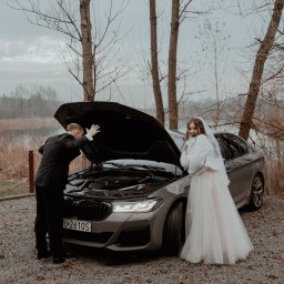 FOCUS4 PRODUCTION Klaudia Karczmarz - Sesja ślubna z humorem: Pan młody zagląda pod maskę szarego BMW, panna młoda w sukni i futerku uśmiecha się obok auta na tle jeziora i drzew bez liści.
