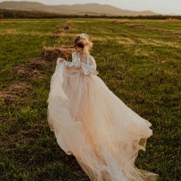 FOCUS4 PRODUCTION Klaudia Karczmarz - Panna młoda w zwiewnej sukni spaceruje po łące o zachodzie słońca. Delikatne światło podkreśla fakturę sukni i naturalny krajobraz w tle.