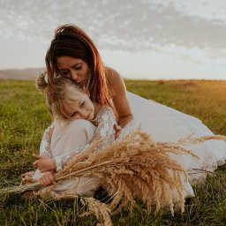 FOCUS4 PRODUCTION Klaudia Karczmarz - Matka w białej sukni obejmuje córkę z bukietem traw na łące o zachodzie słońca. Ciepłe światło, naturalna sceneria, intymna chwila.