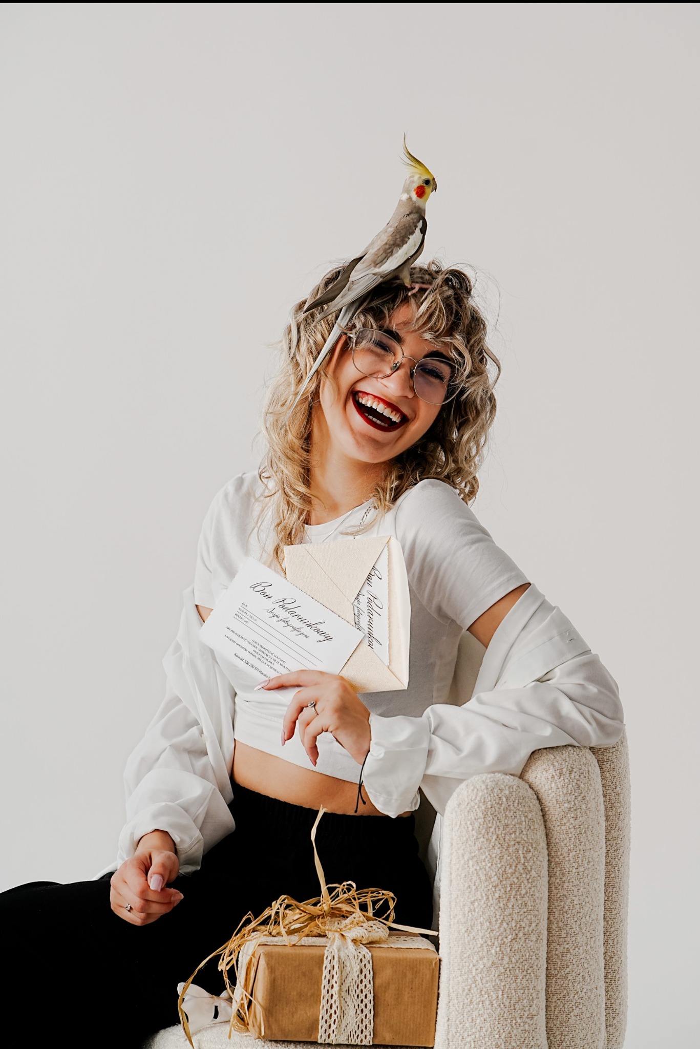 Uśmiechnięta kobieta w okularach trzyma bon podarunkowy na sesję fotograficzną, na głowie siedzi papuga. Prezent zapakowany obok na fotelu.