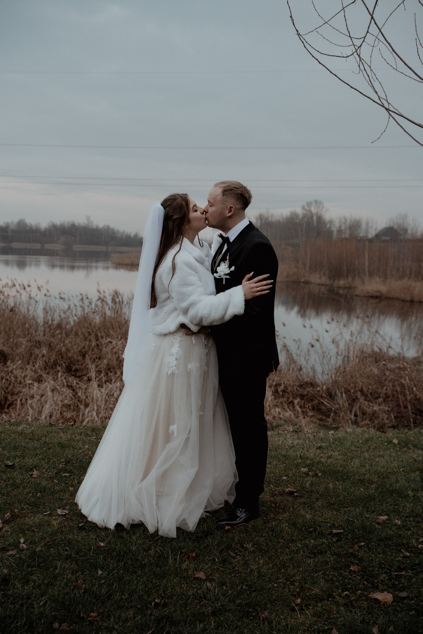 Pocałunek pary młodej w plenerze nad jeziorem, panna młoda w białym futerku i sukni z koronkowymi aplikacjami, pan młody w czarnym garniturze.