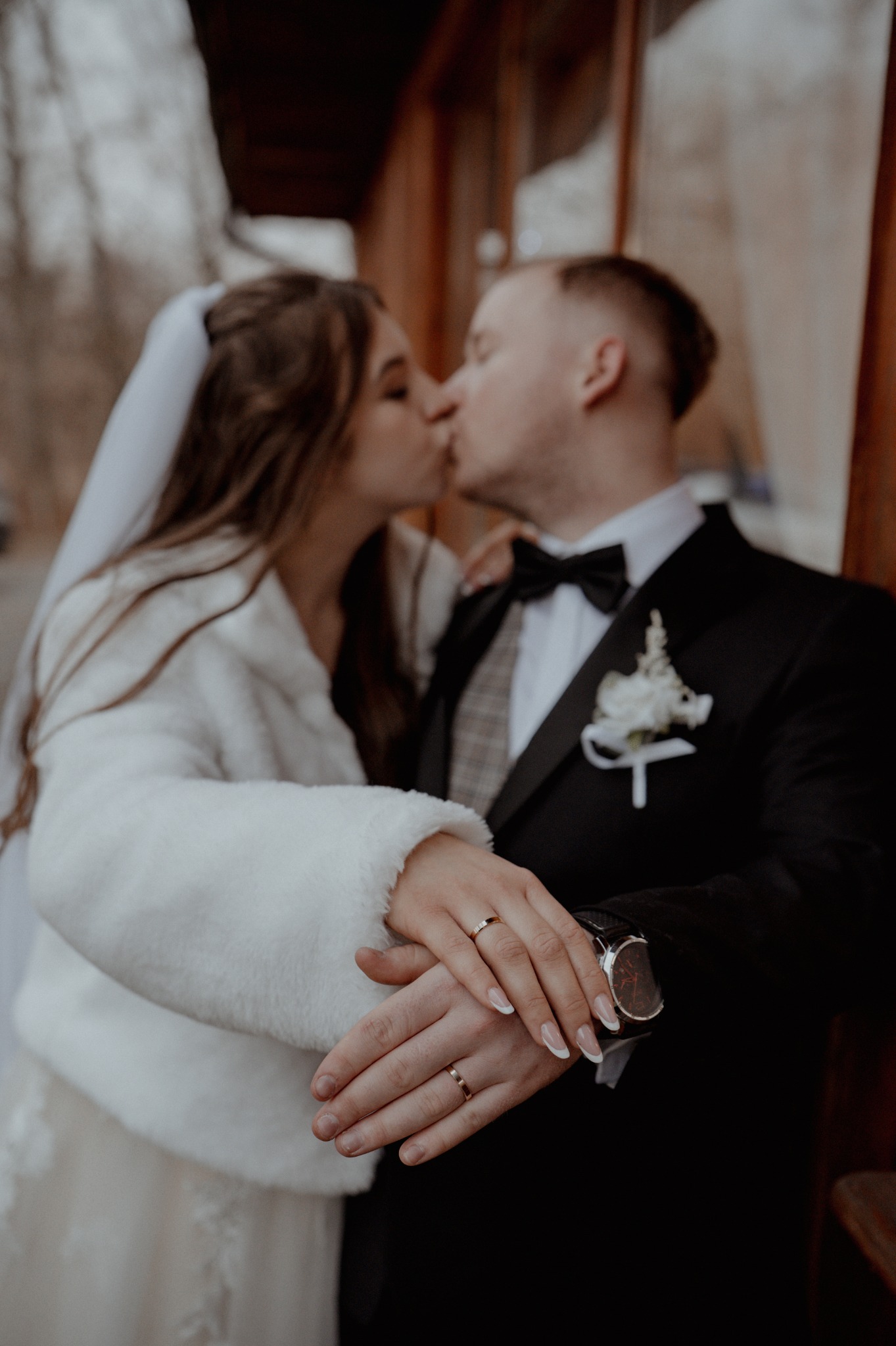 Pocałunek nowożeńców ze złotymi obrączkami w Świebodzicach. Panna młoda w białym futerku, pan młody w czarnym smokingu z muchą i kwiatem.