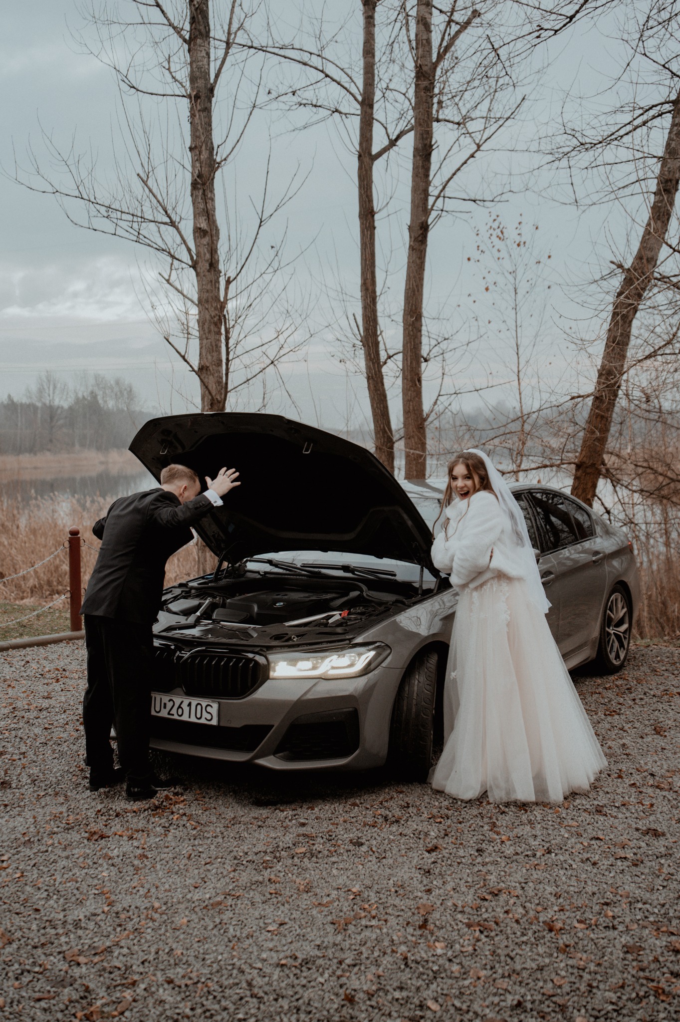 Sesja ślubna z humorem: Pan młody zagląda pod maskę szarego BMW, panna młoda w sukni i futerku uśmiecha się obok auta na tle jeziora i drzew bez liści.