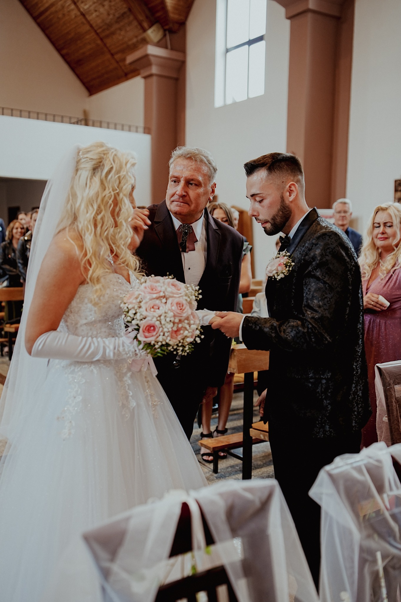 Uroczystość ślubna w kościele: panna młoda w białej sukni i welonie trzyma bukiet róż, obok pan młody w czarnym fraku, w tle goście i ojciec panny młodej.