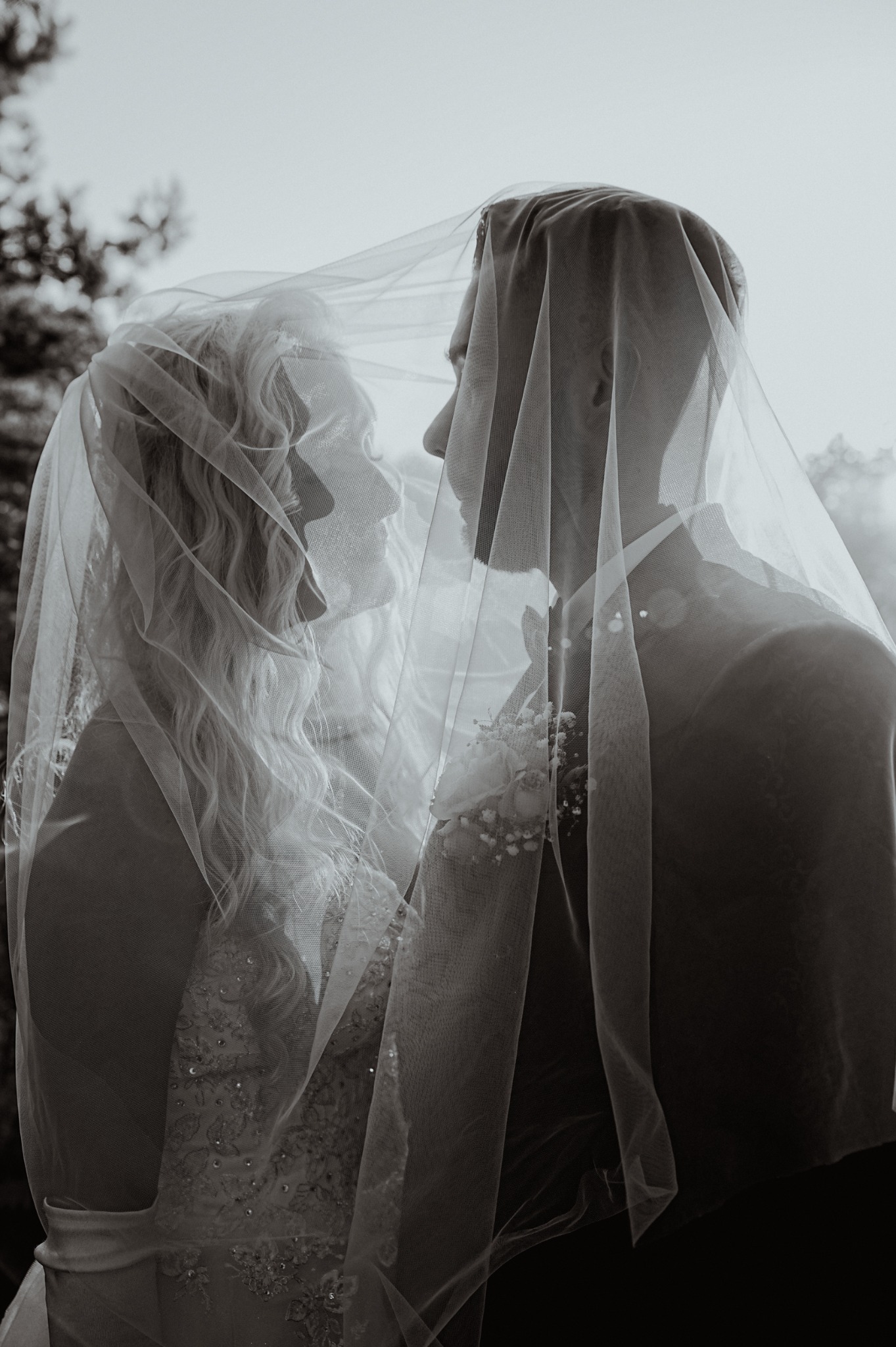 Czarno-białe zbliżenie pary młodej pod welonem, zarys sylwetek na tle rozmytej zieleni, delikatne światło podkreśla intymność chwili.