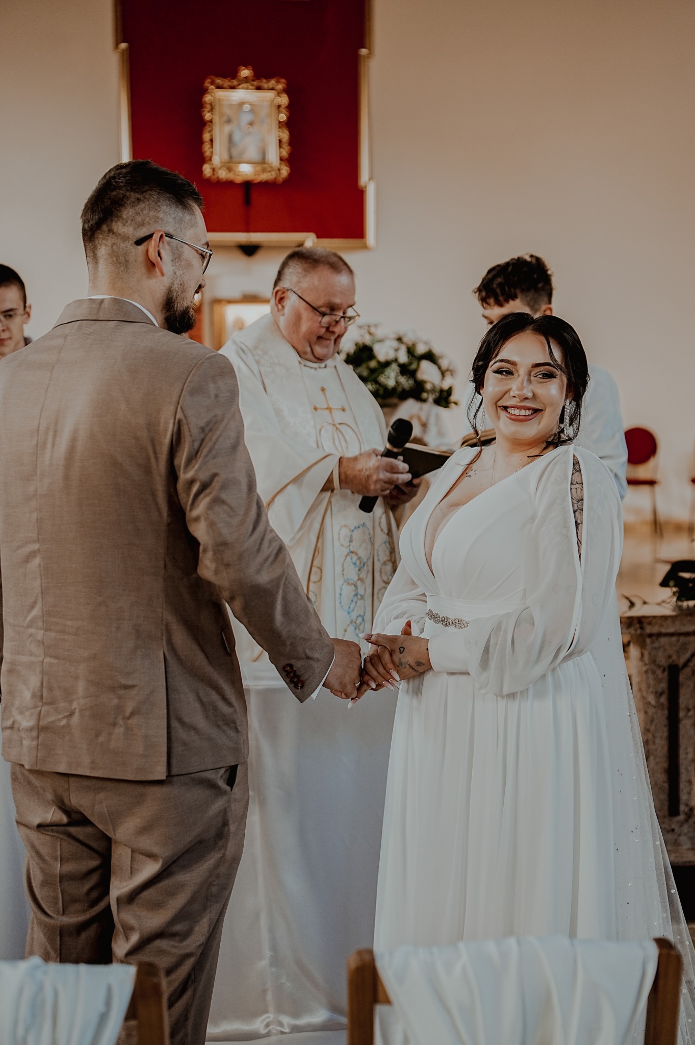 Uśmiechnięta panna młoda w białej sukni, trzymająca dłoń pana młodego podczas ceremonii ślubnej w kościele, z księdzem w tle i dekoracją kwiatową.