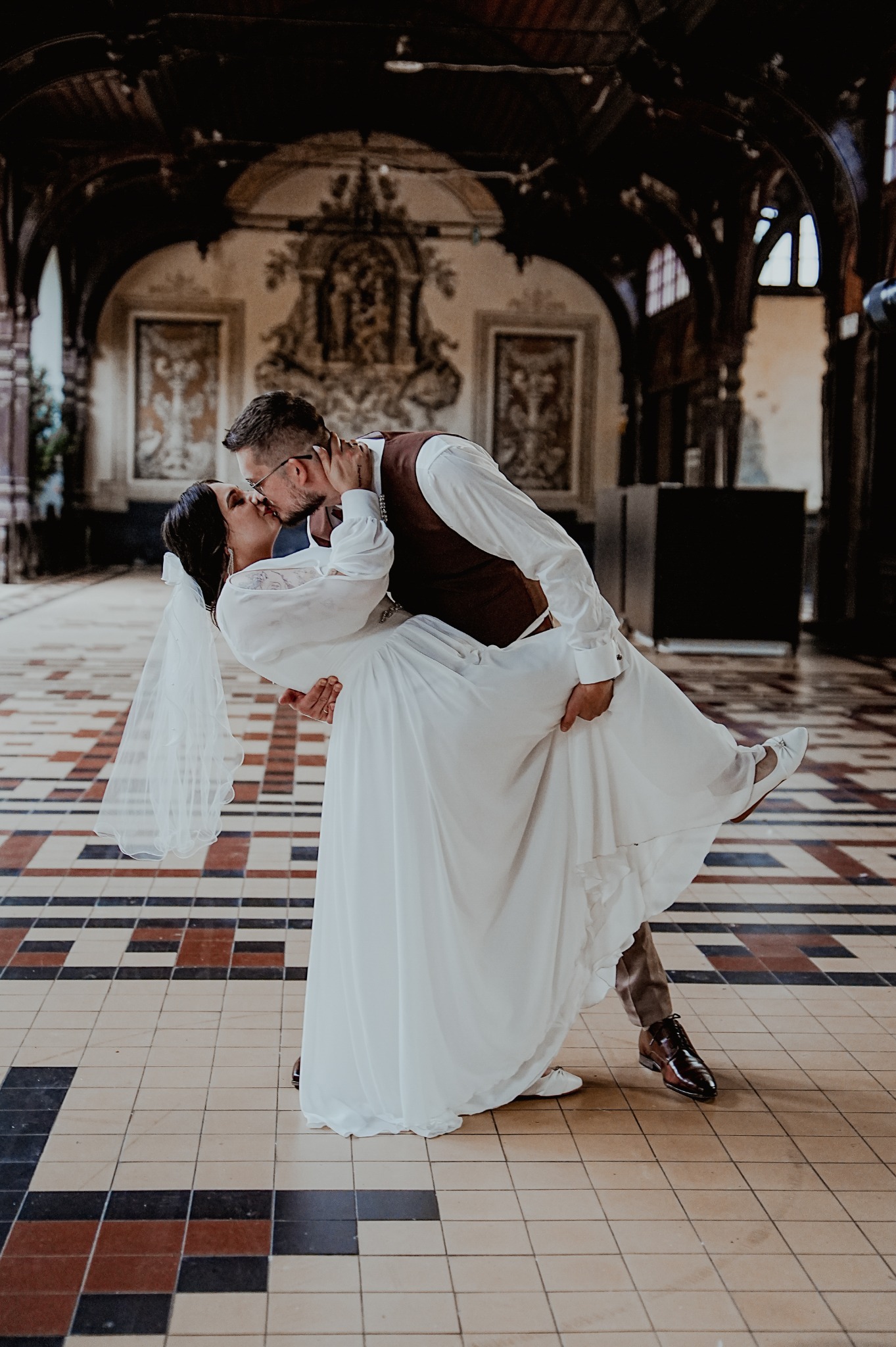 Panna młoda w białej sukni i welonie, unoszona przez pana młodego w brązowej kamizelce, całują się na tle dekoracyjnego wnętrza z wzorzystą podłogą.