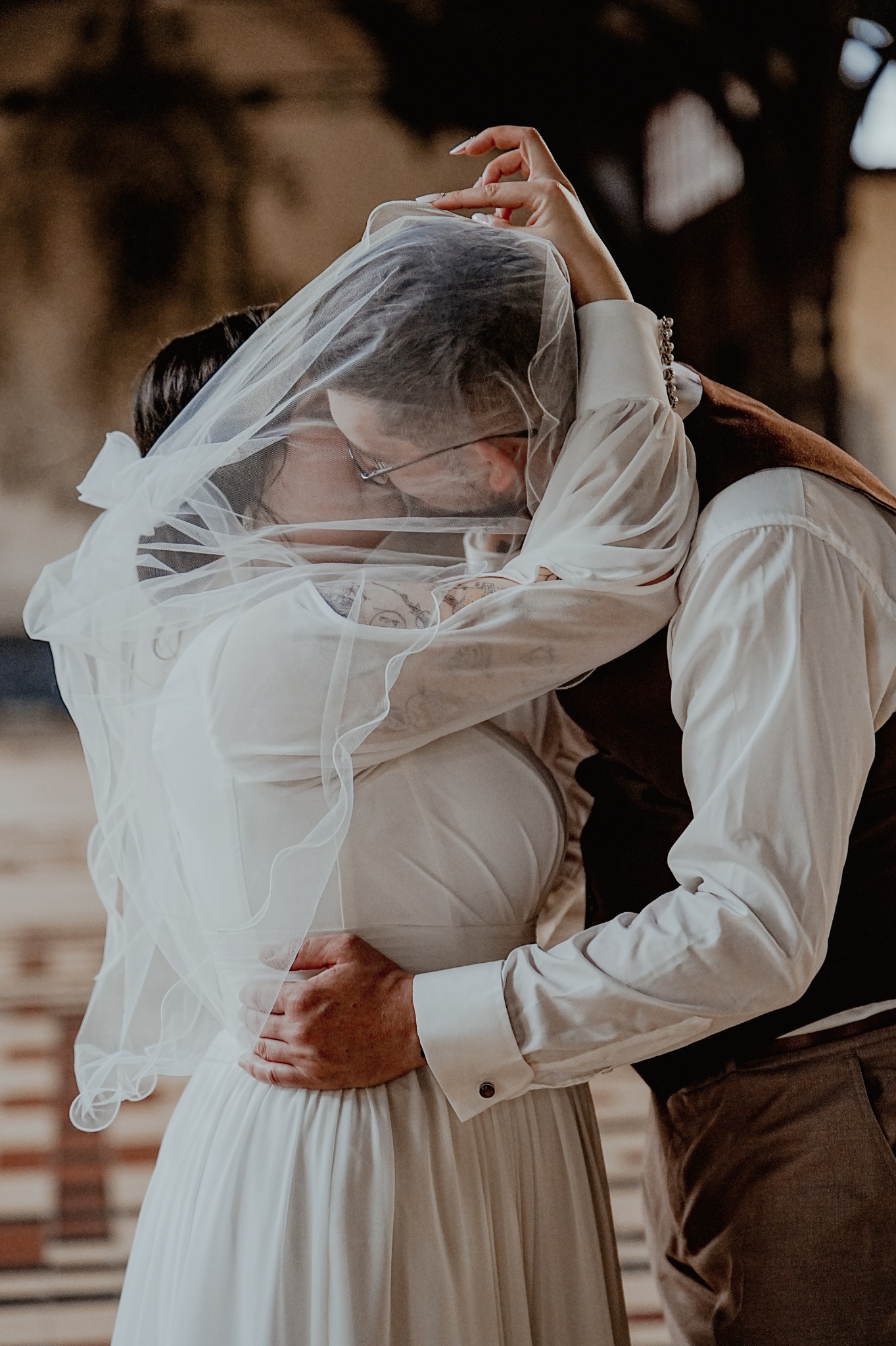 Pocałunek pary młodej pod welonem. Pan młody w okularach i tatuażem na ramieniu obejmuje pannę młodą w białej sukni. Delikatne światło podkreśla intymność chwili.