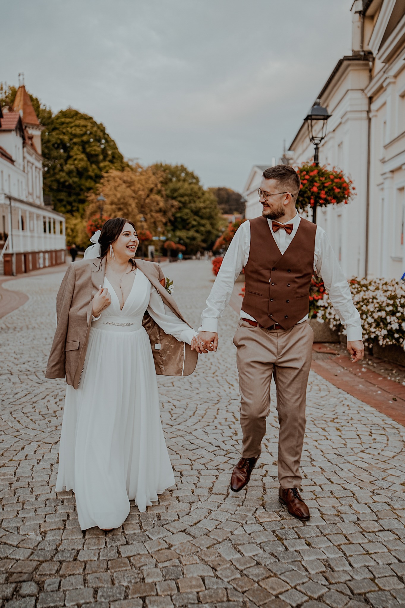 Uśmiechnięta para młoda, idąca po brukowanej ulicy, trzymając się za ręce. Ona w sukni ślubnej z marynarką, on w kamizelce i muszce. Kwiaty w tle.