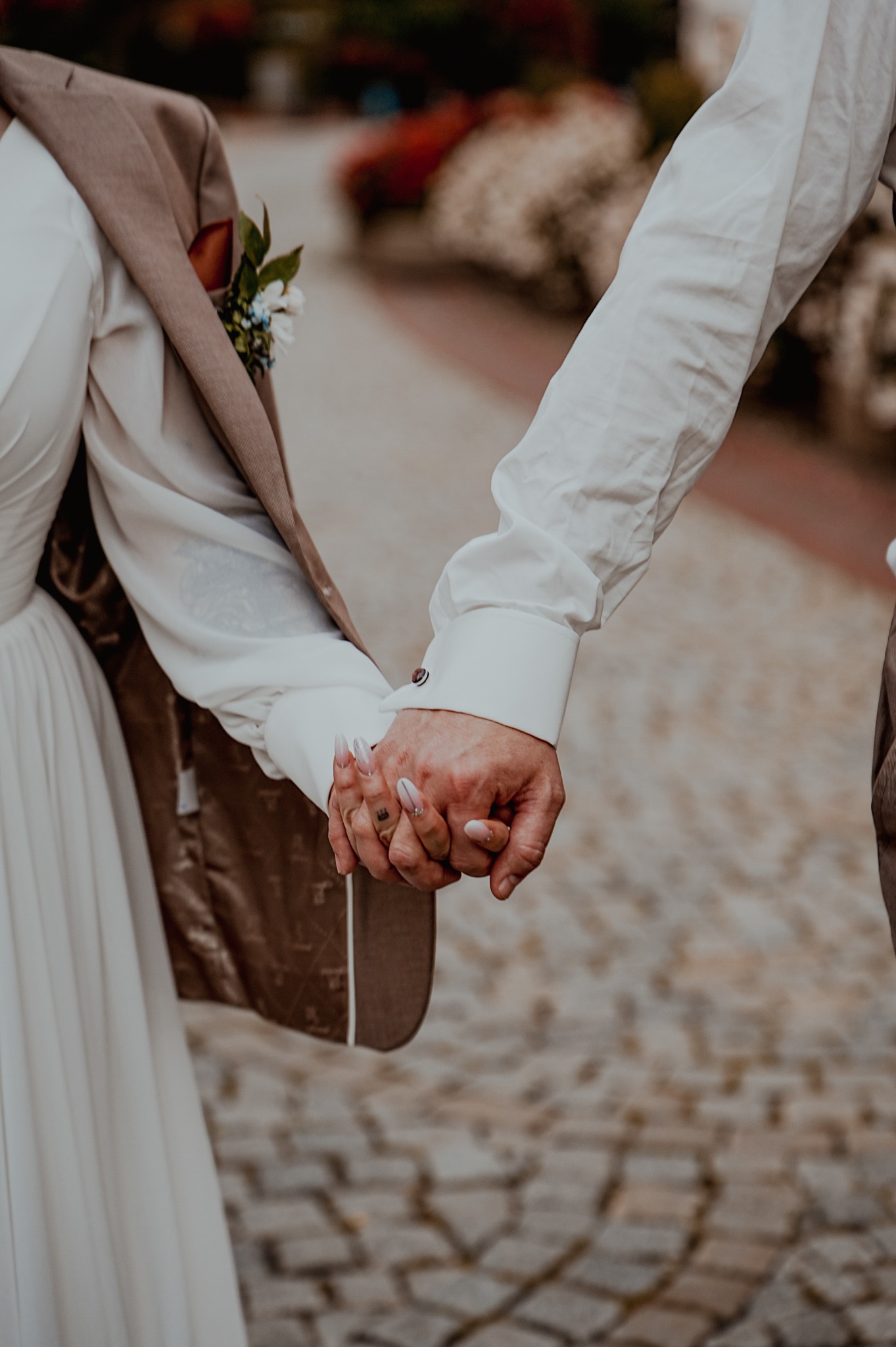 Zbliżenie dłoni pary młodej trzymających się na tle brukowanej ścieżki. Ona w białej sukni, on w koszuli z mankietami i brązowym garniturze z butonierką.