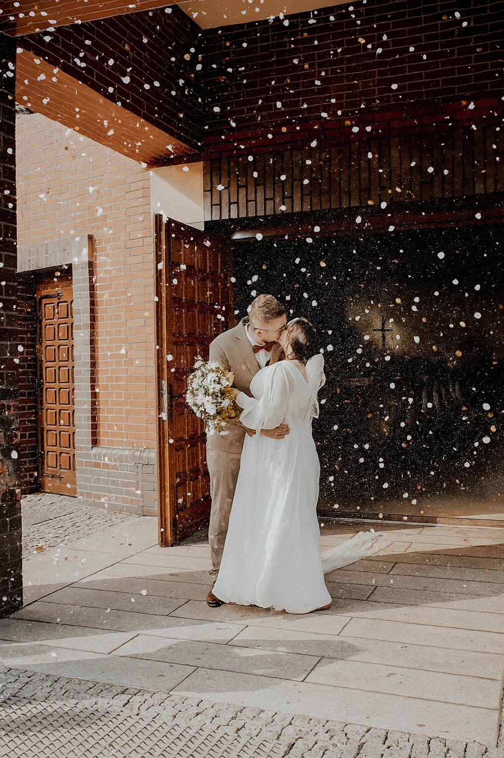 Pocałunek nowożeńców przed kościołem, obsypanych konfetti. Panna młoda w białej sukni, pan młody w beżowym garniturze z muchą. Ujęcie w ciepłych barwach.