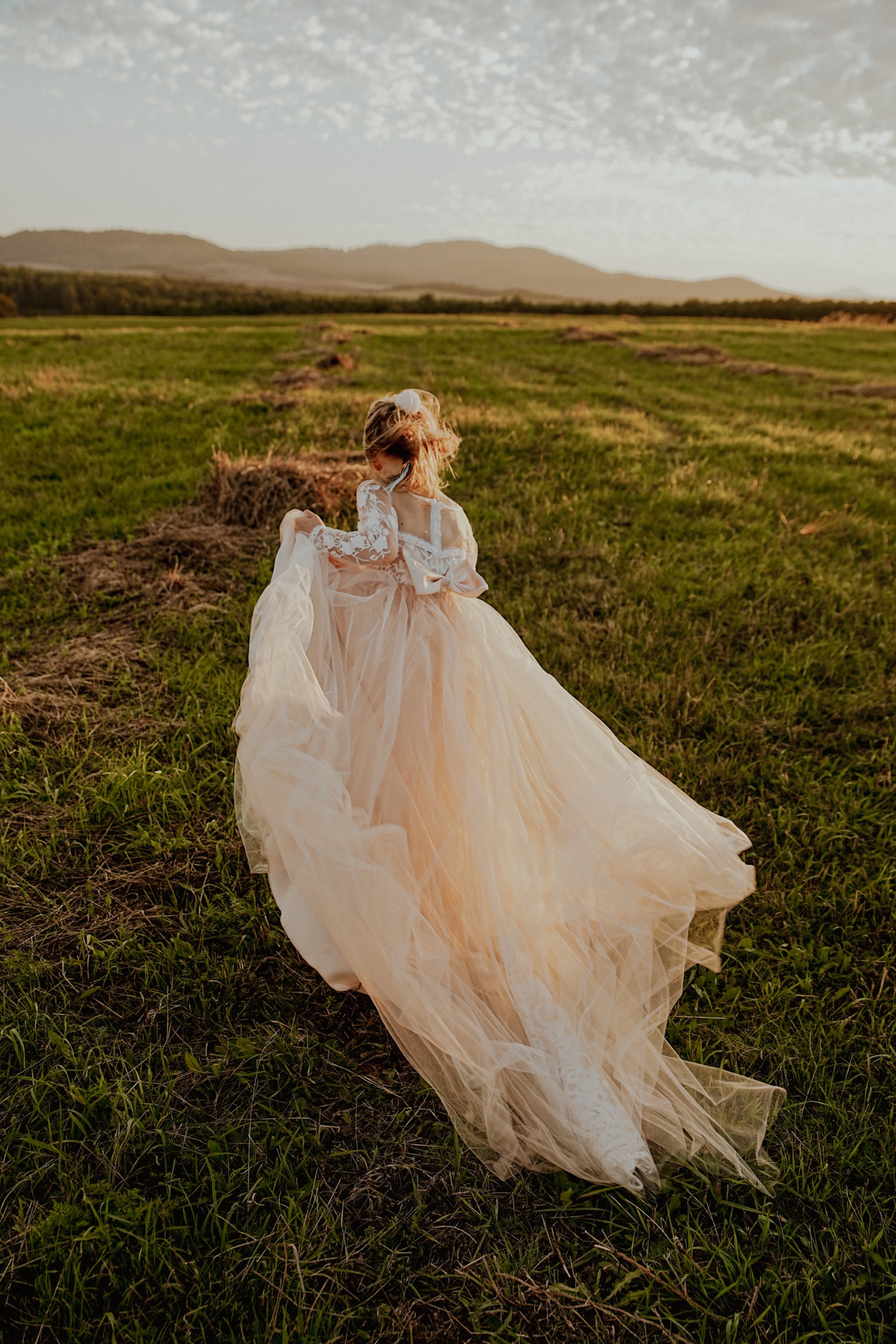 Panna młoda w zwiewnej sukni spaceruje po łące o zachodzie słońca. Delikatne światło podkreśla fakturę sukni i naturalny krajobraz w tle.