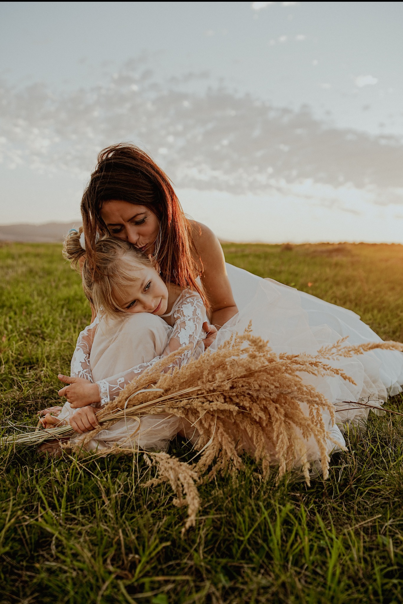 Matka w białej sukni obejmuje córkę z bukietem traw na łące o zachodzie słońca. Ciepłe światło, naturalna sceneria, intymna chwila.