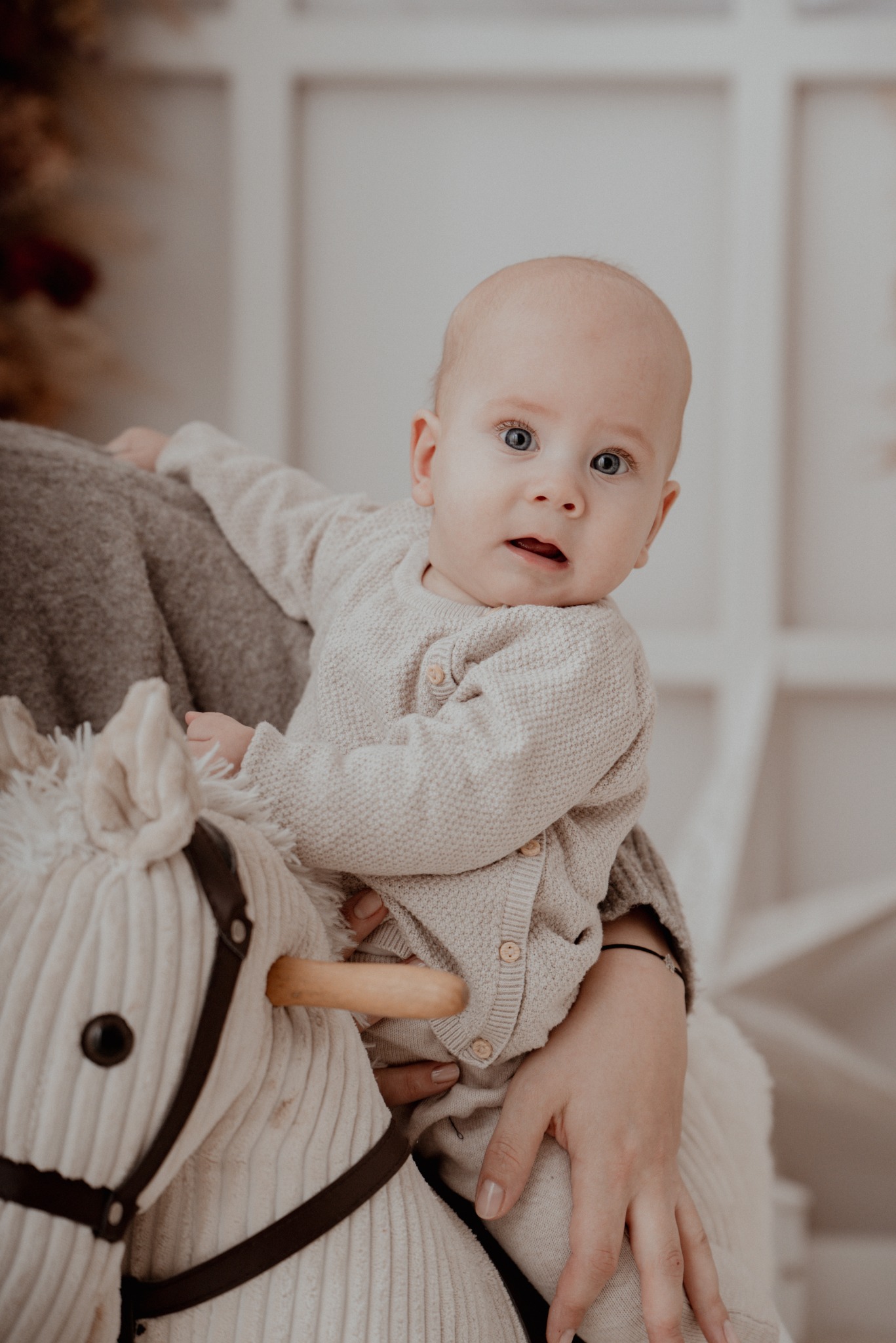 Niemowlę w beżowym sweterku siedzi na pluszowym koniku, trzymane przez osobę w szarym swetrze. Jasne tło z elementami dekoracyjnymi.