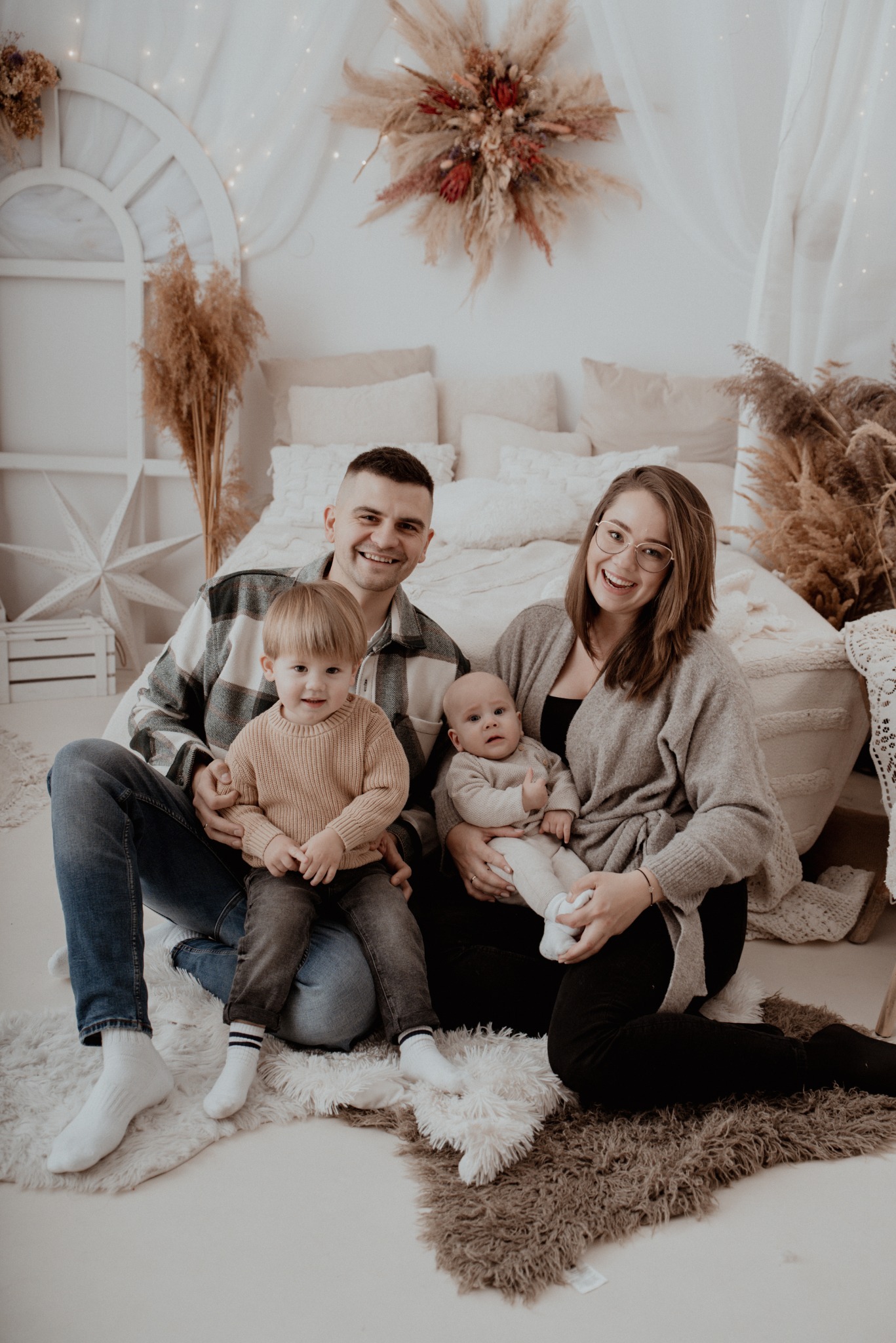 Uśmiechnięta rodzina z dwójką małych dzieci siedzi na podłodze w jasnym, studyjnym wnętrzu z dekoracjami z suszonych traw i łóżkiem w tle. Ciepła, naturalna kolorystyka.