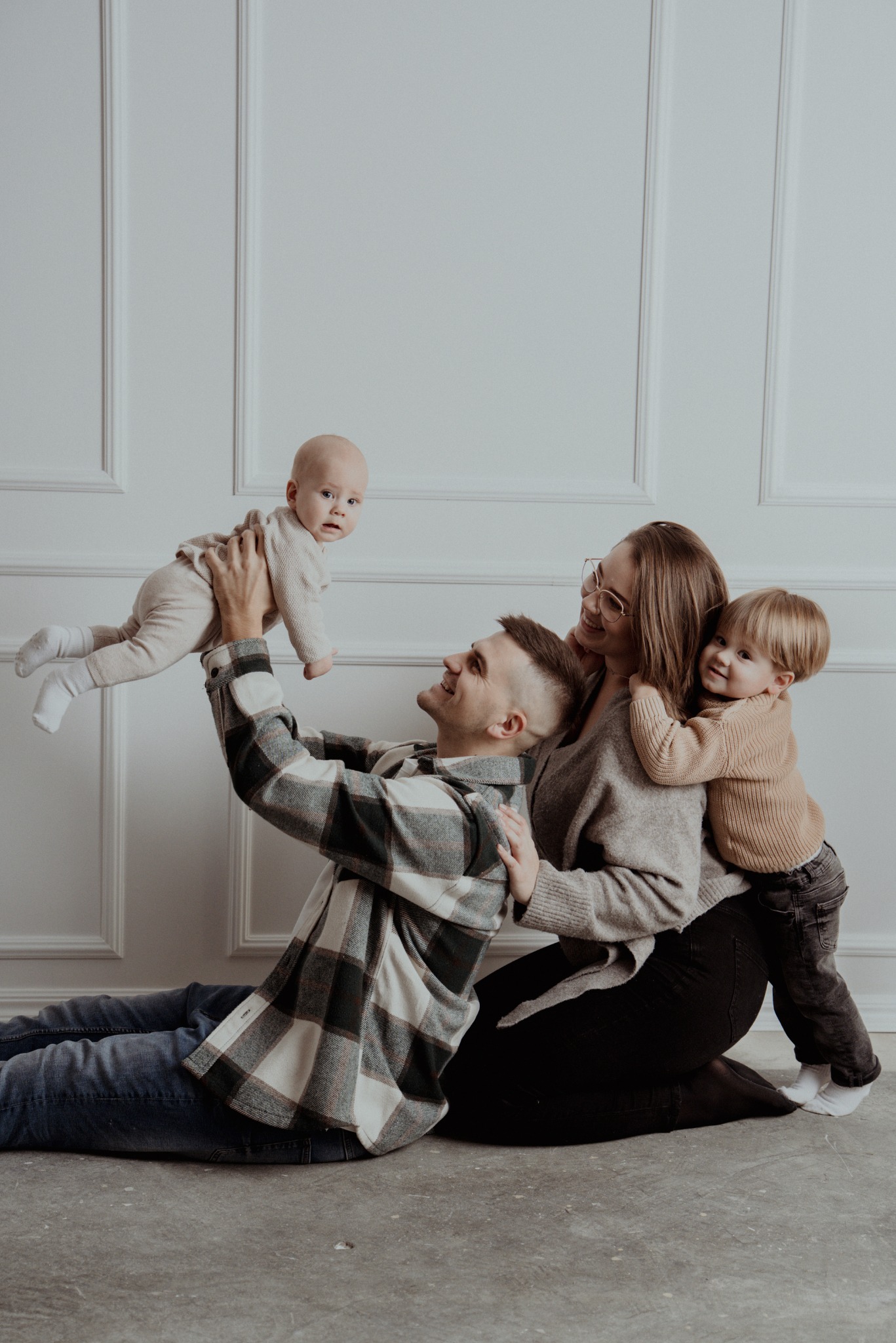 Rodzinne ujęcie w studio: tata podrzuca niemowlę, mama z synkiem przytulają się, jasne tło z panelami, naturalne światło, ciepła kolorystyka, swobodna atmosfera.