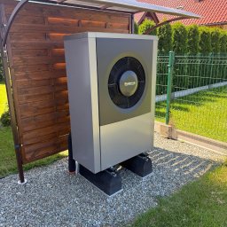 INSTAL-HEAT KRZYSZTOF RUSINEK - Szara pompa ciepła Buderus na żwirowym podłożu, osłonięta drewnianą konstrukcją z przezroczystym zadaszeniem, na tle zielonego trawnika i ogrodzenia.