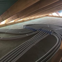 INSTAL-HEAT KRZYSZTOF RUSINEK - Szare rury instalacji ogrzewania podłogowego, ułożone równolegle na szarej powierzchni, z drewnianą konstrukcją dachu w tle. Widok z góry, szeroki kąt.