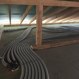 INSTAL-HEAT KRZYSZTOF RUSINEK - Szare rury ogrzewania podłogowego ułożone równolegle na szarym podłożu, widoczne drewniane elementy konstrukcji dachu, instalacja przed wylaniem posadzki.