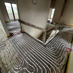 INSTAL-HEAT KRZYSZTOF RUSINEK - Rozłożona instalacja ogrzewania podłogowego w surowym wnętrzu domu, widoczne białe rury na folii z czerwoną siatką, żółta taśma przy ścianach, schody w tle.