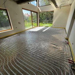 INSTAL-HEAT KRZYSZTOF RUSINEK - Białe rury ogrzewania podłogowego ułożone na folii w surowym wnętrzu z dużymi oknami i betonowym stropem. Widoczne elementy instalacji przy ścianach.