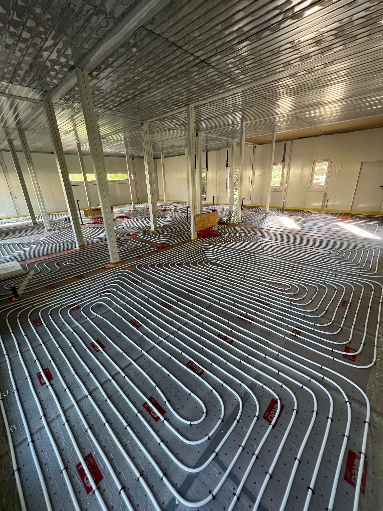 Białe rury ogrzewania podłogowego ułożone spiralnie na szarej macie w przestronnym, surowym wnętrzu z metalowym sufitem i białymi ścianami, widoczne słupy podtrzymujące.