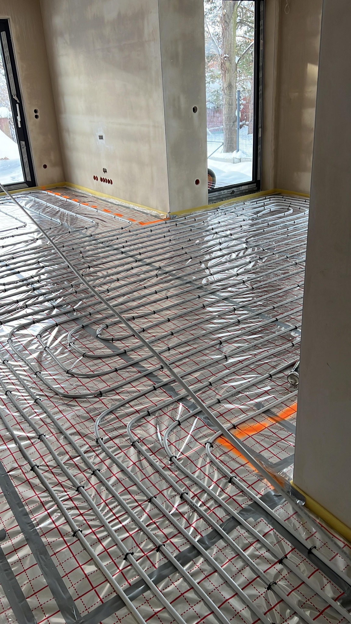 Ułożone rury ogrzewania podłogowego na folii izolacyjnej w surowym wnętrzu z oknem. Widoczne mocowania rur i taśma zabezpieczająca ściany.