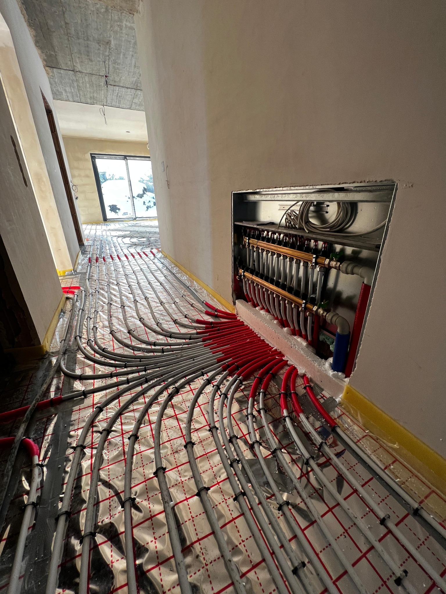 Rozłożona instalacja ogrzewania podłogowego z rozdzielaczem w ścianie, widok z góry, rury w kolorze szarym i czerwonym na folii z czerwoną kratką, surowy stan wykończenia.