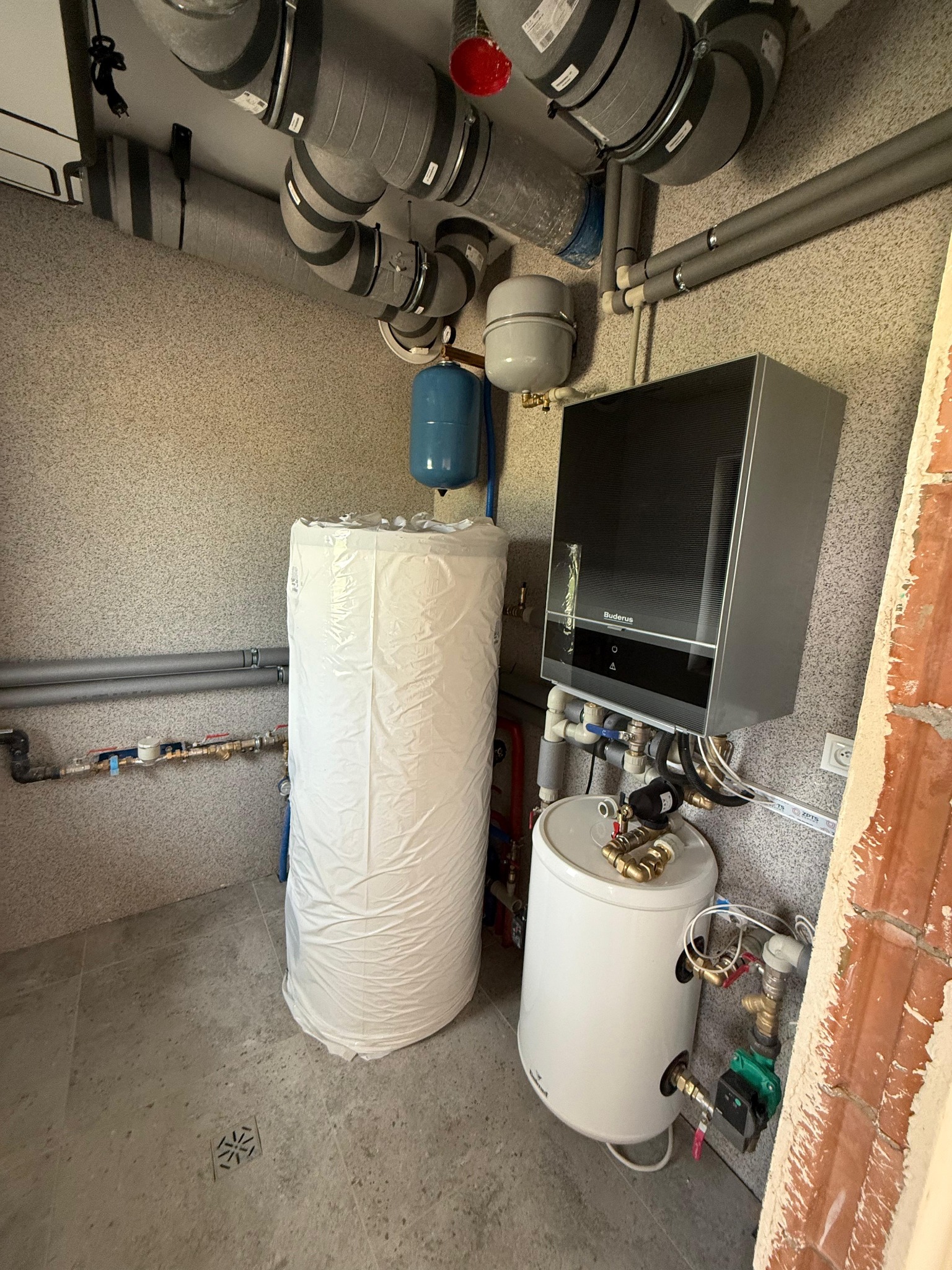 Zestaw grzewczy Buderus z zasobnikiem ciepłej wody, zbiornikiem wyrównawczym i rozbudowaną instalacją wentylacyjną w pomieszczeniu technicznym. Widoczne rury i zawory.