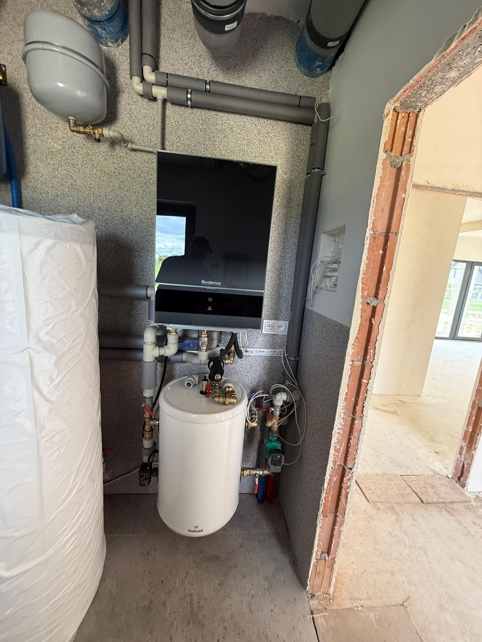 Nowoczesny piec gazowy Buderus z białym zasobnikiem ciepłej wody, zamontowany na ścianie w pomieszczeniu w trakcie wykańczania, widoczne rury i instalacje.