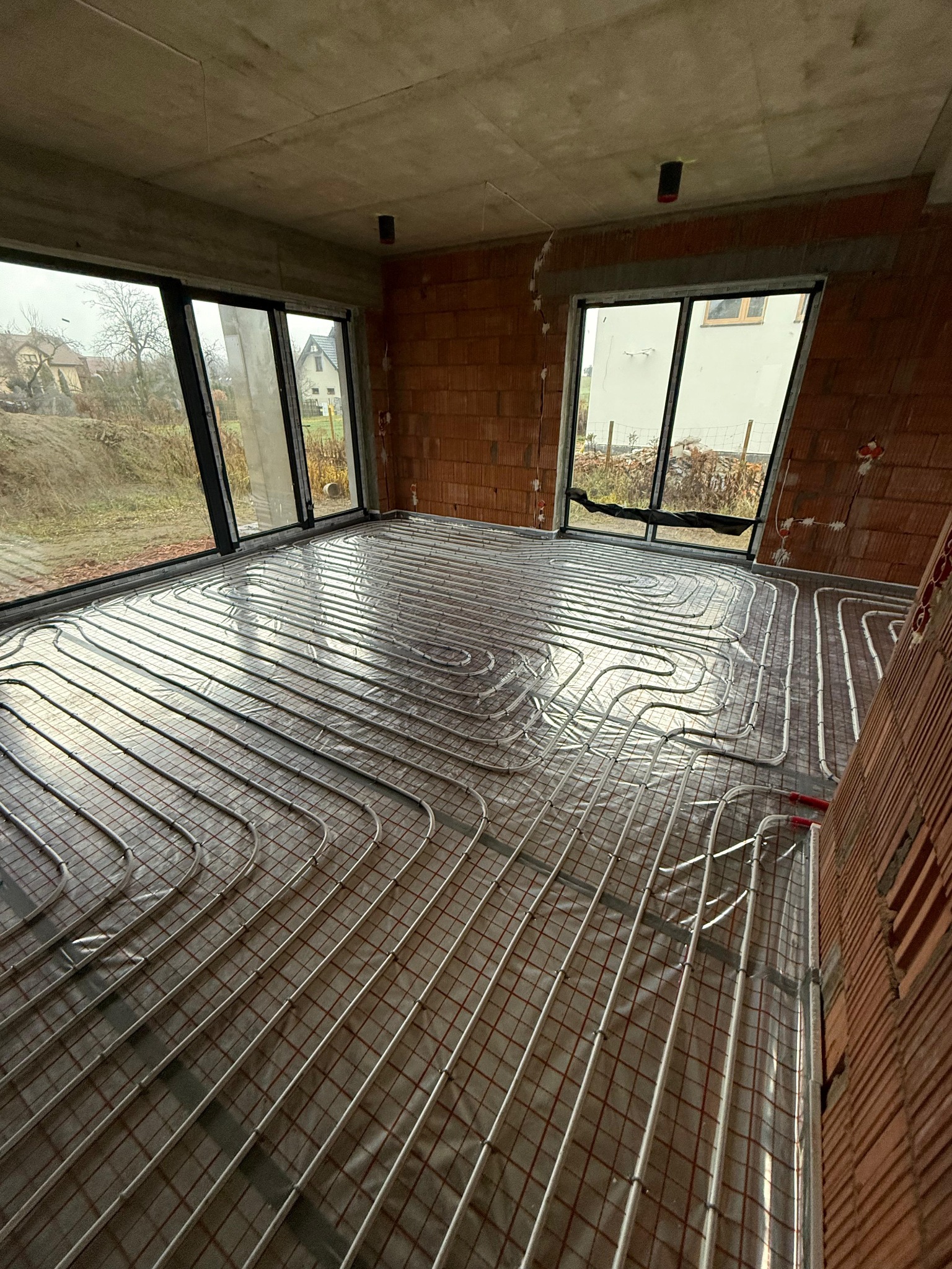 Instalacja ogrzewania podłogowego w surowym wnętrzu z ceglanymi ścianami i dużymi oknami. Widoczne rury grzewcze ułożone na izolacji.